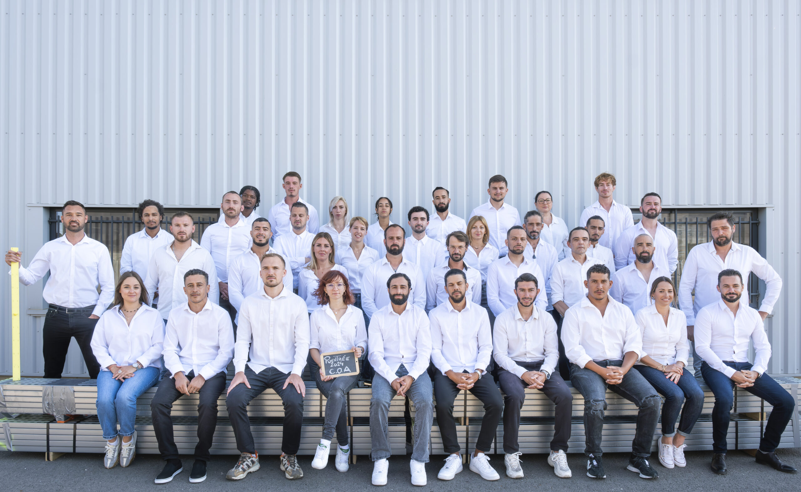 Grand groupe de trente-cinq collaborateurs en chemises blanches posant sur des marches devant un bâtiment industriel, alignés.