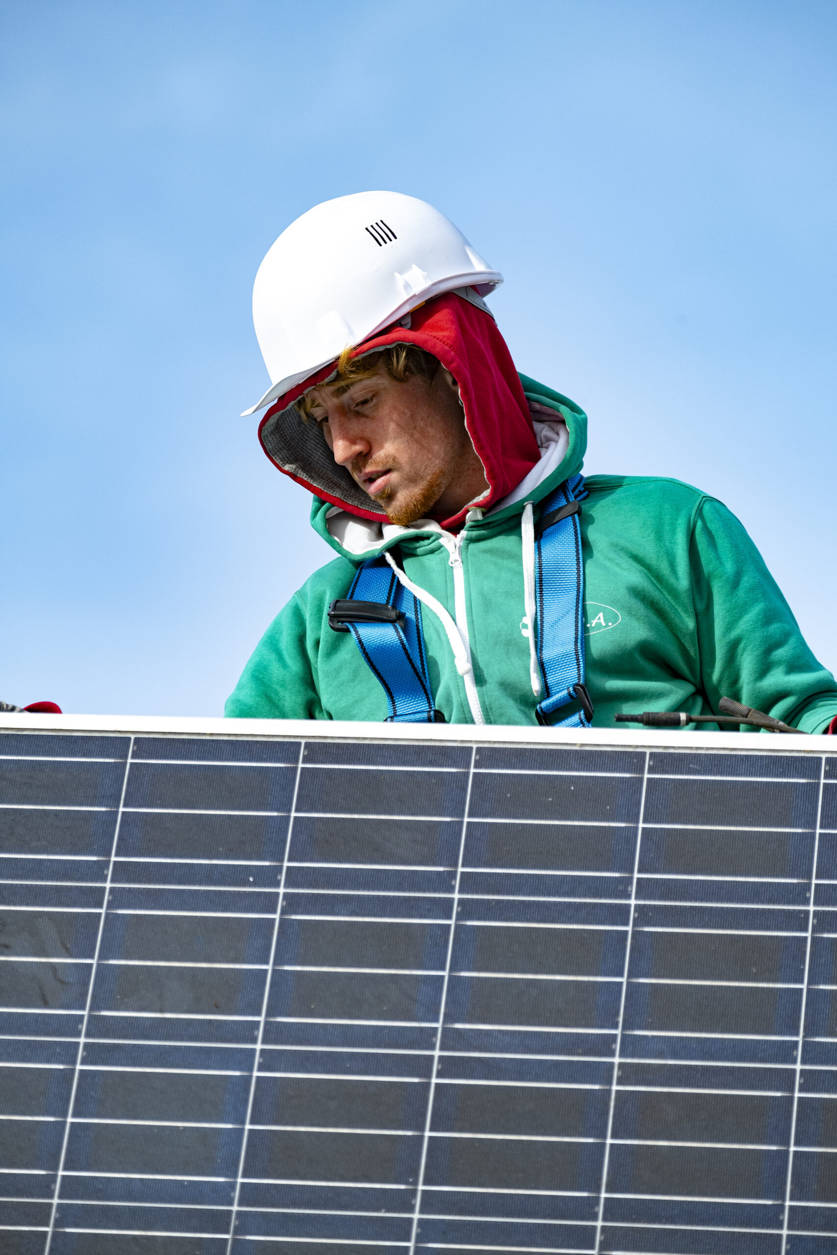 Technicien en casque et harnais posant un panneau solaire, sweat vert C.O.A., ciel bleu en arrière-plan.