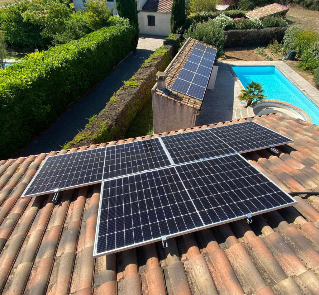 Vue en légère plongée de nouveaux modules solaires bleus fixés sur une toiture en tuiles canal, à côté d’une piscine et d’un pool house déjà équipé de panneaux en arrière-plan.