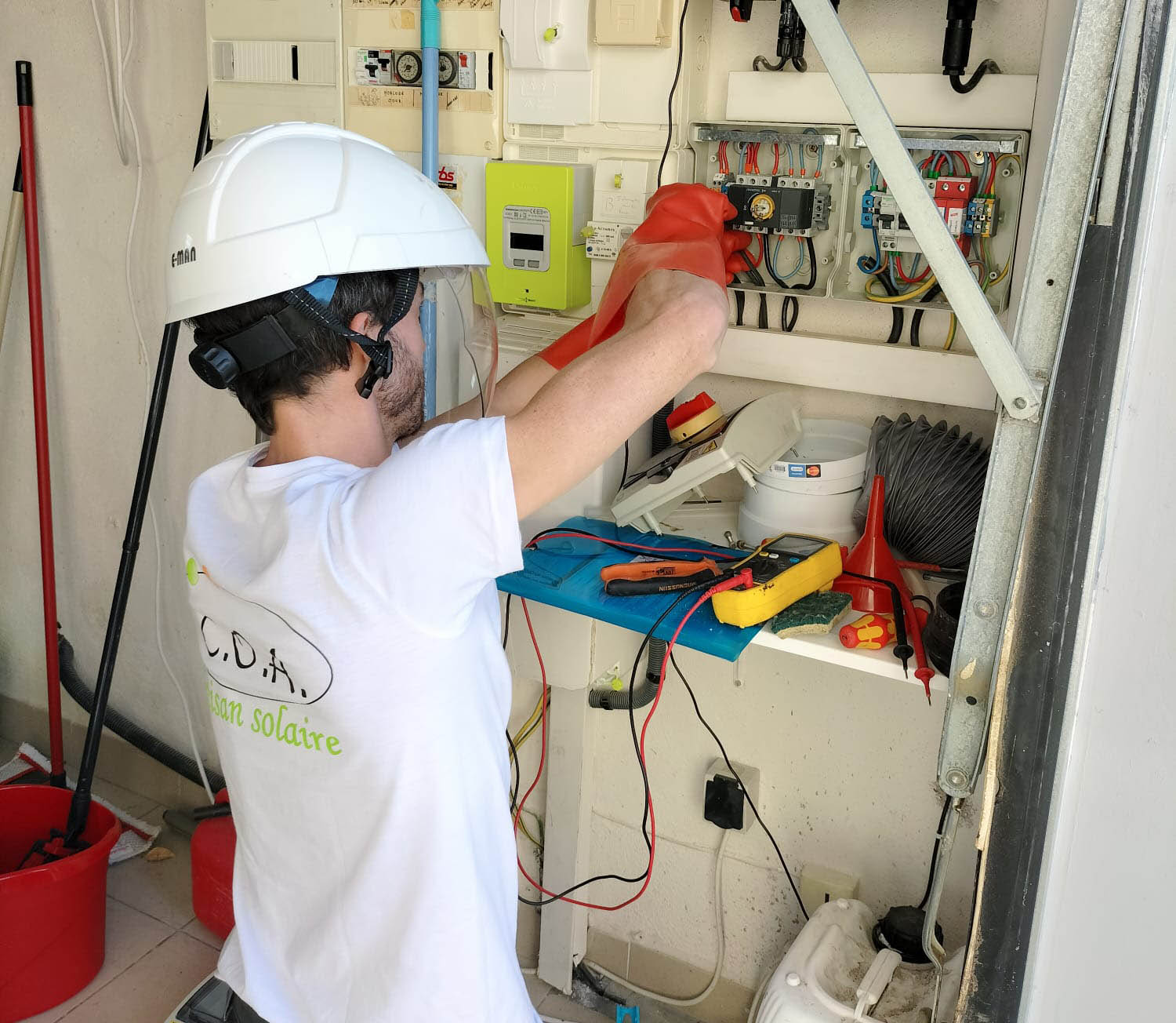 Installateur en t-shirt C.O.A et casque de protection, ganté, travaillant sur les connexions d’un onduleur solaire vert dans un coffret mural avec câbles et outils de mesure posés à portée de main.