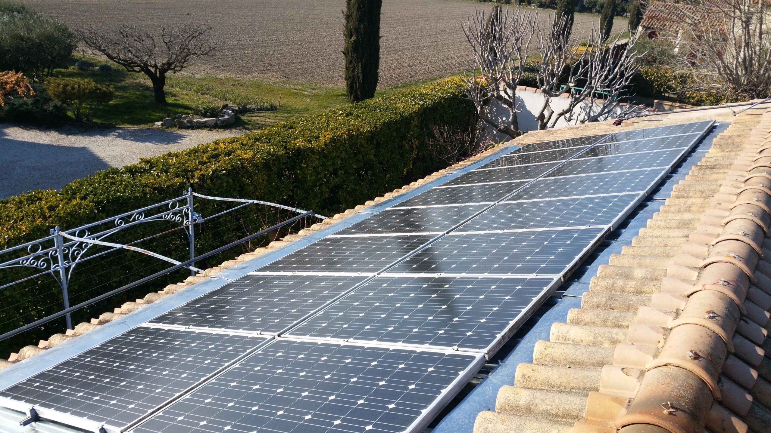 Vue oblique de modules solaires bleus encastrés et affleurants dans une toiture en tuiles terre cuite, munis de solins métalliques, avec une haie, un champ et des arbres en arrière-plan.