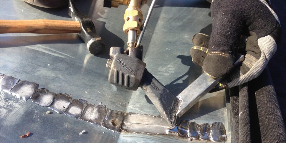 Vue rapprochée d’une main gantée manipulant un chalumeau et un fer à souder pour fondre de l’étain sur un joint en tôle, avec un marteau posé à côté.