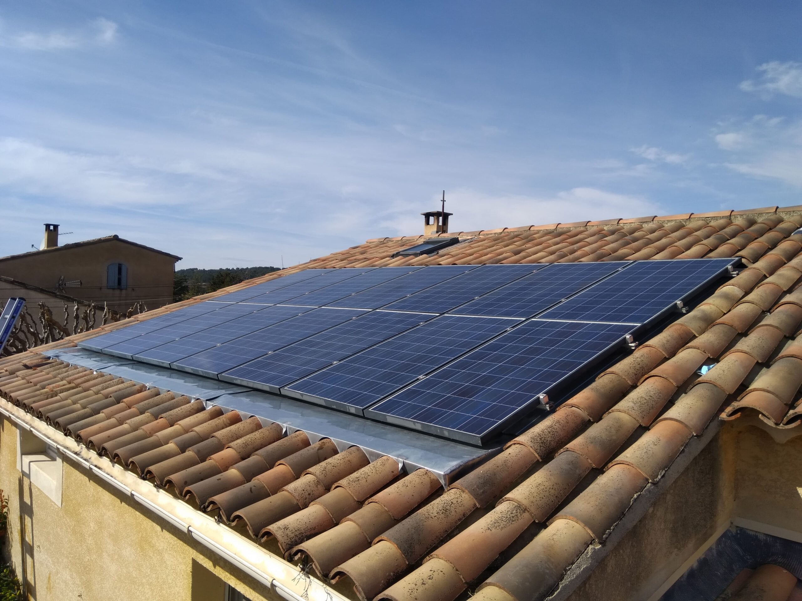 Vue oblique de modules solaires bleus encastrés et affleurants dans une toiture en tuiles canal terre cuite, avec solins métalliques visibles.