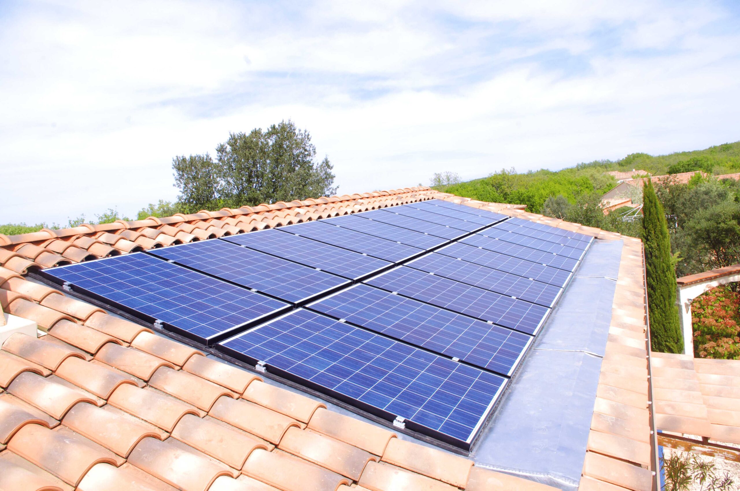 Vue en angle de panneaux solaires photovoltaïques bleus intégrés dans une toiture en tuiles terre cuite, alignés et affleurant les tuiles, avec un arrière-plan de végétation