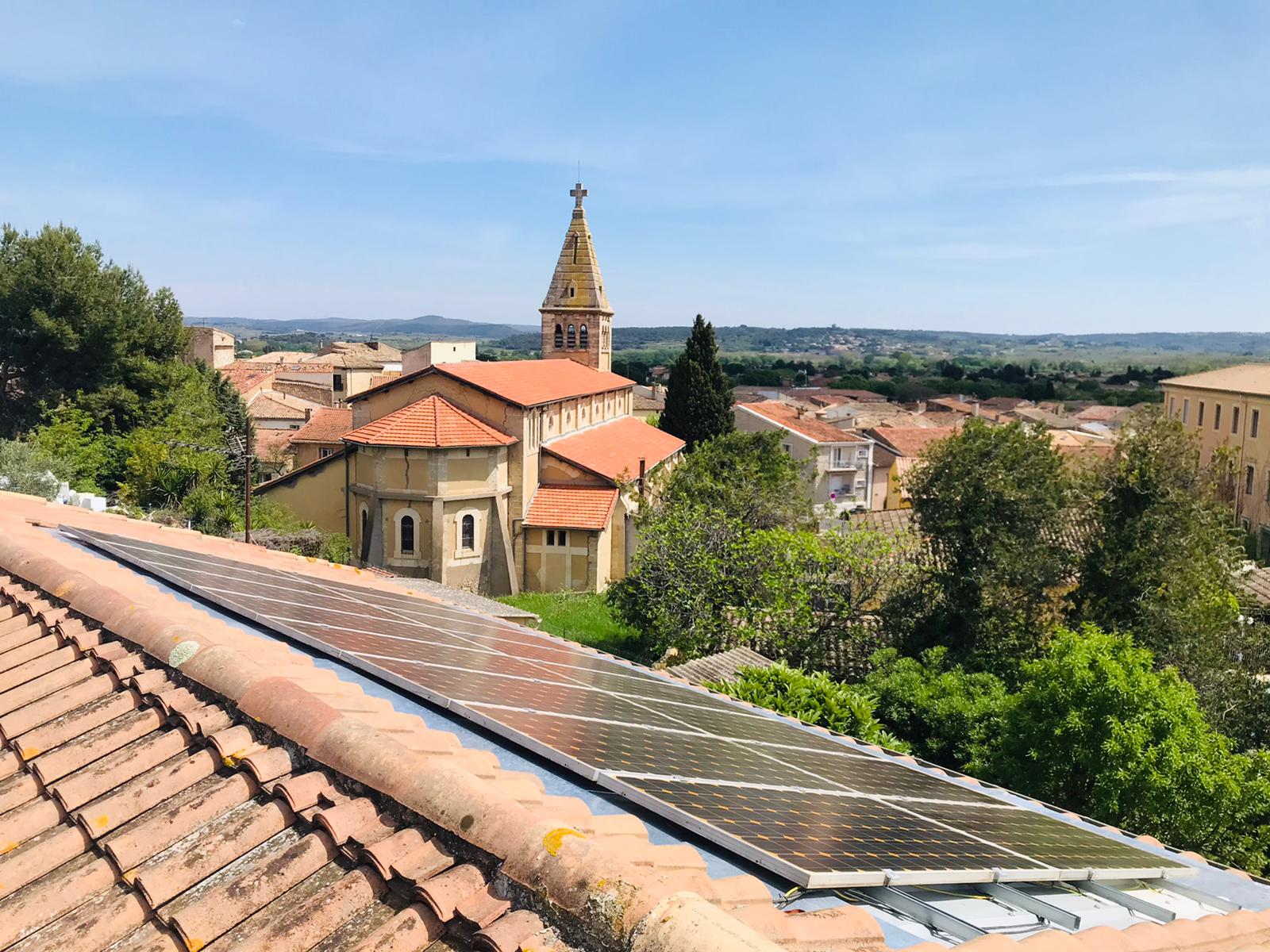 Vue oblique de modules solaires bleus encastrés dans une toiture en tuiles terre cuite, avec en arrière-plan un clocher d’église, d’autres toits et un paysage verdoyant.