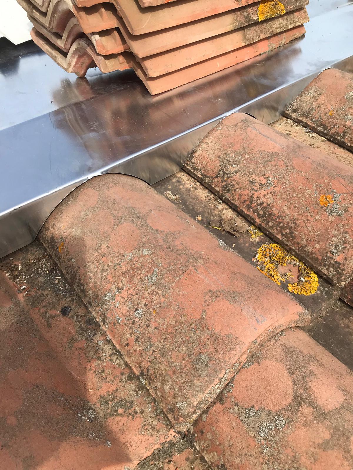 Gros plan sur un bac d'étanchéité en zinc pliée brillante installé entre deux rangées de tuiles canal terre cuite, avec un tas de tuiles empilées en arrière-plan.