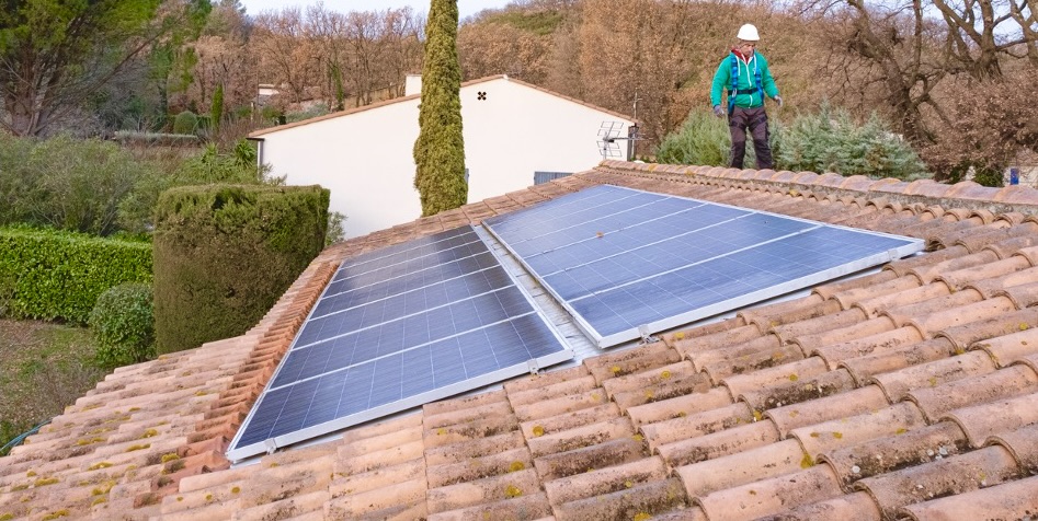Toit en tuiles avec deux rangées de panneaux photovoltaïques et un technicien en casque vérifiant l'installation.
