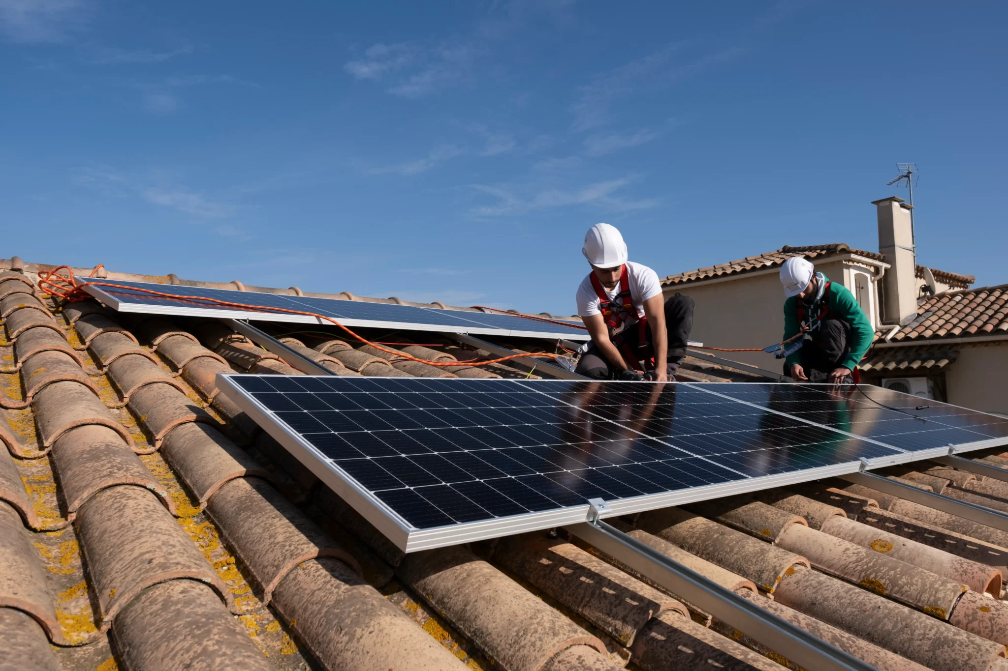 Deux techniciens en équipement de sécurité (casques blancs et harnais) ajustent et fixent des panneaux solaires noirs sur une toiture en tuiles canal, avec rails de montage et cordages d’ancrage visibles, sous un ciel bleu.