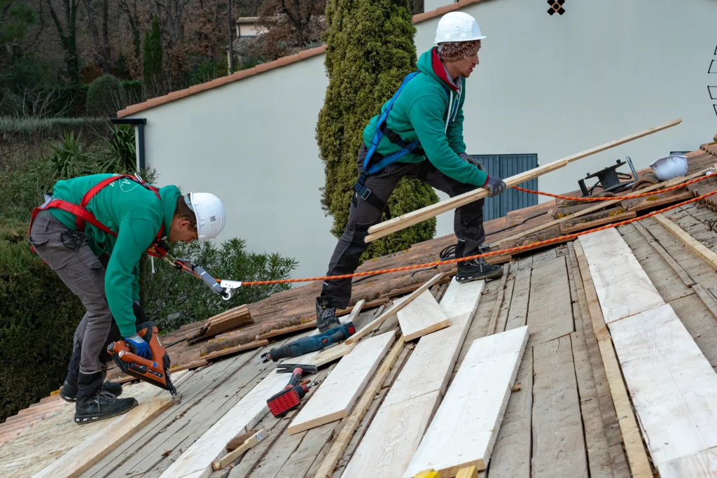 Deux ouvriers en casques blancs et harnais bleus et rouges travaillent sur une toiture : l’un cloue des lattes de bois, l’autre apporte une planche, avec outils et câbles de sécurité à leurs pieds.