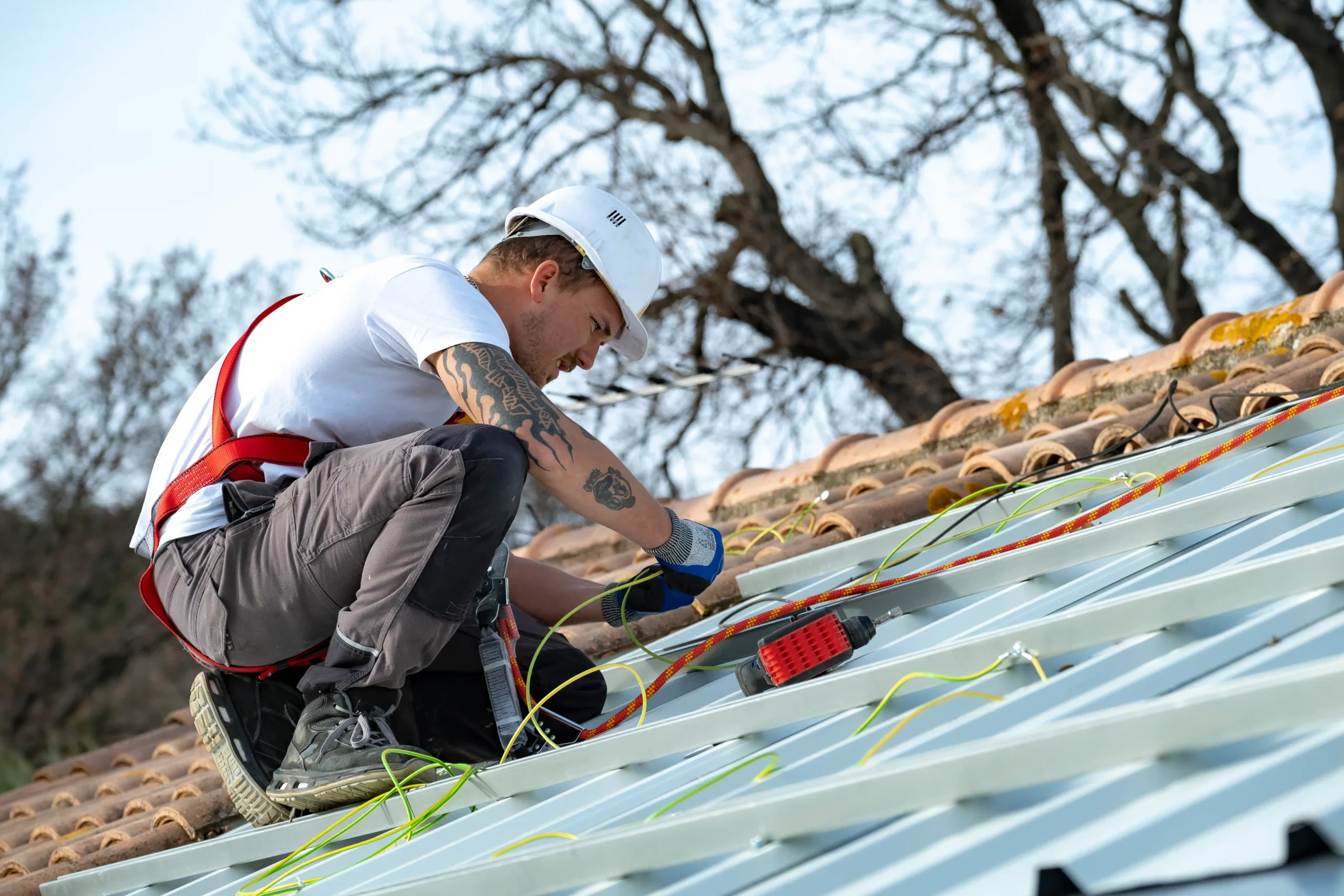 Homme en casque de chantier et harnais rouge agenouillé sur une toiture métallique, connectant des câbles verts et jaunes à la structure de fixation d’un panneau solaire.