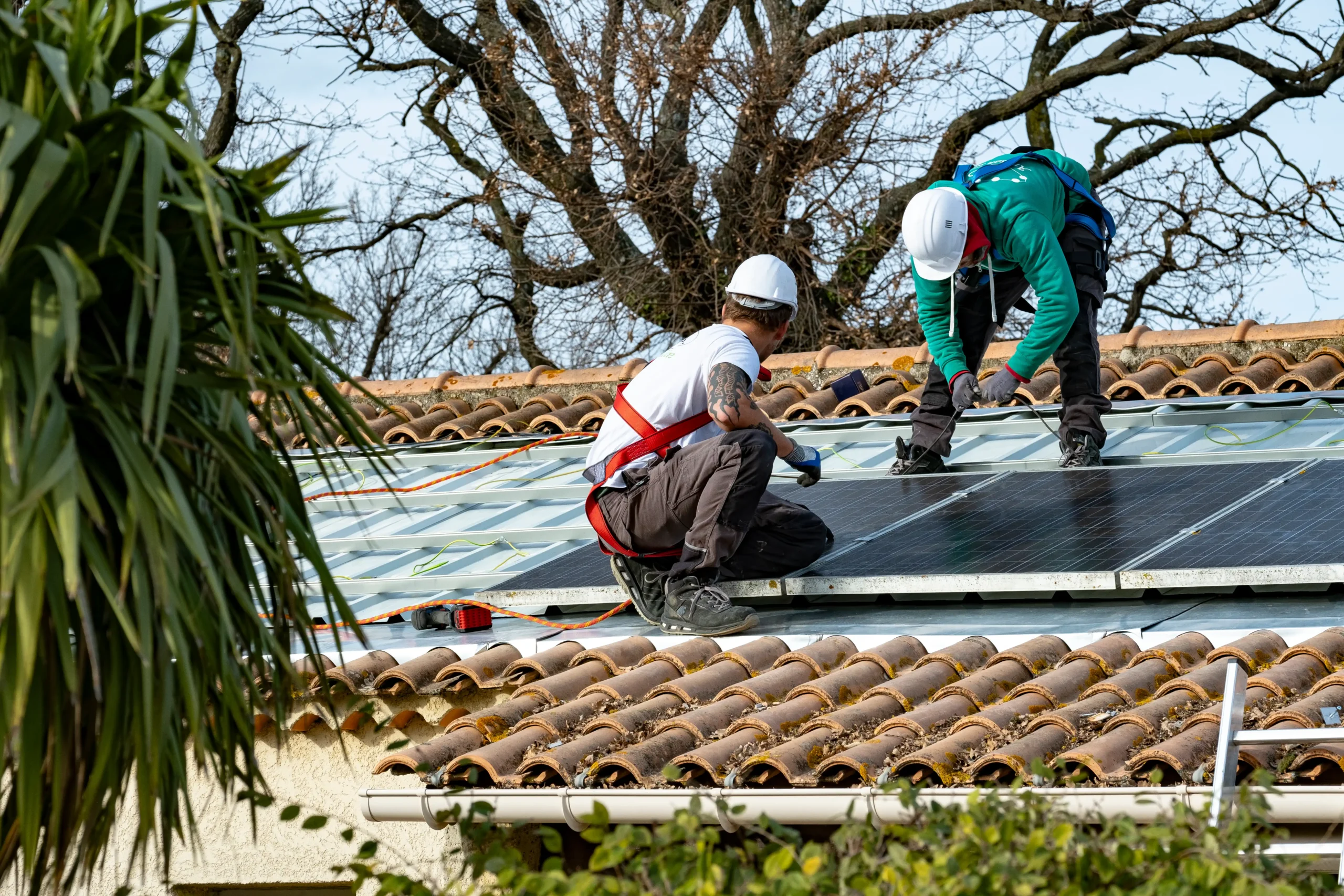 Deux techniciens en tenue de chantier (casques, harnais et gants) entretiennent des panneaux solaires noirs sur une structure bac acier intégrée à une toiture en tuiles canal, avec cordages de sécurité et feuillage en bordure.
