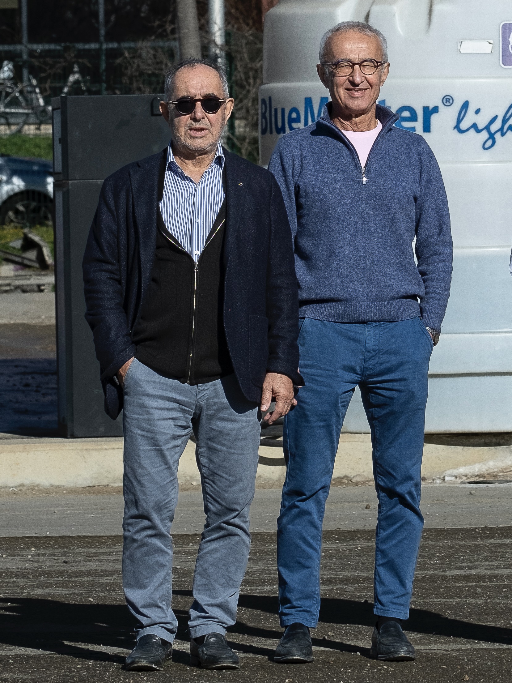 Deux hommes âgés debout côte à côte devant un réservoir, vêtements décontractés, l'un porte des lunettes de soleil.