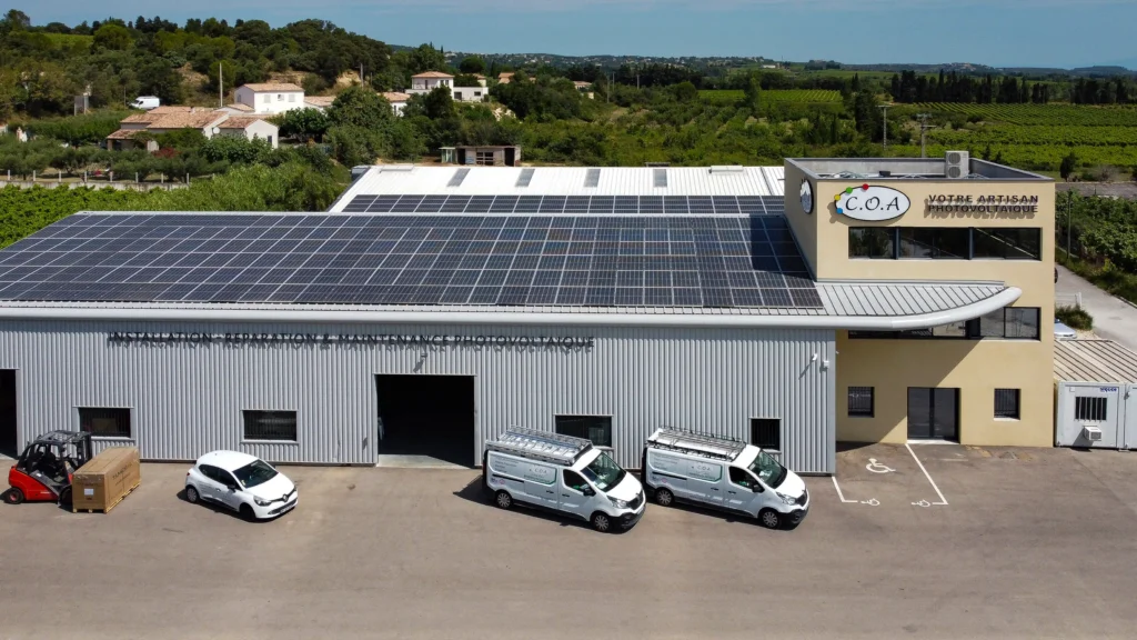 Bâtiment industriel vu en plongée, couvert de panneaux solaires sur son toit en métal, avec trois camionnettes de l’artisan solaire garées devant et un paysage de vignes en arrière-plan.