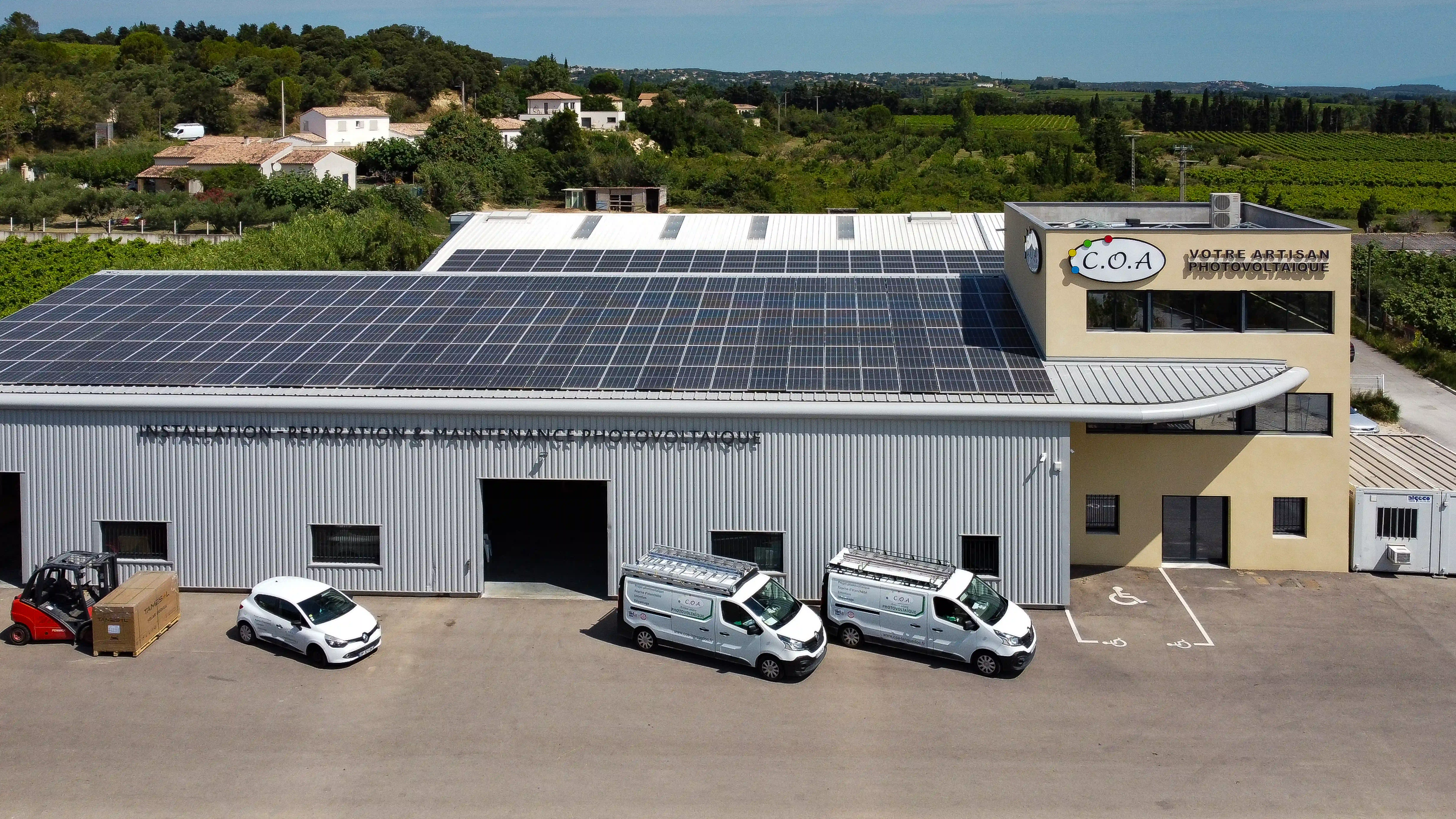 Bâtiment industriel vu en plongée, couvert de panneaux solaires sur son toit en métal, avec trois camionnettes de l’artisan solaire garées devant et un paysage de vignes en arrière-plan.