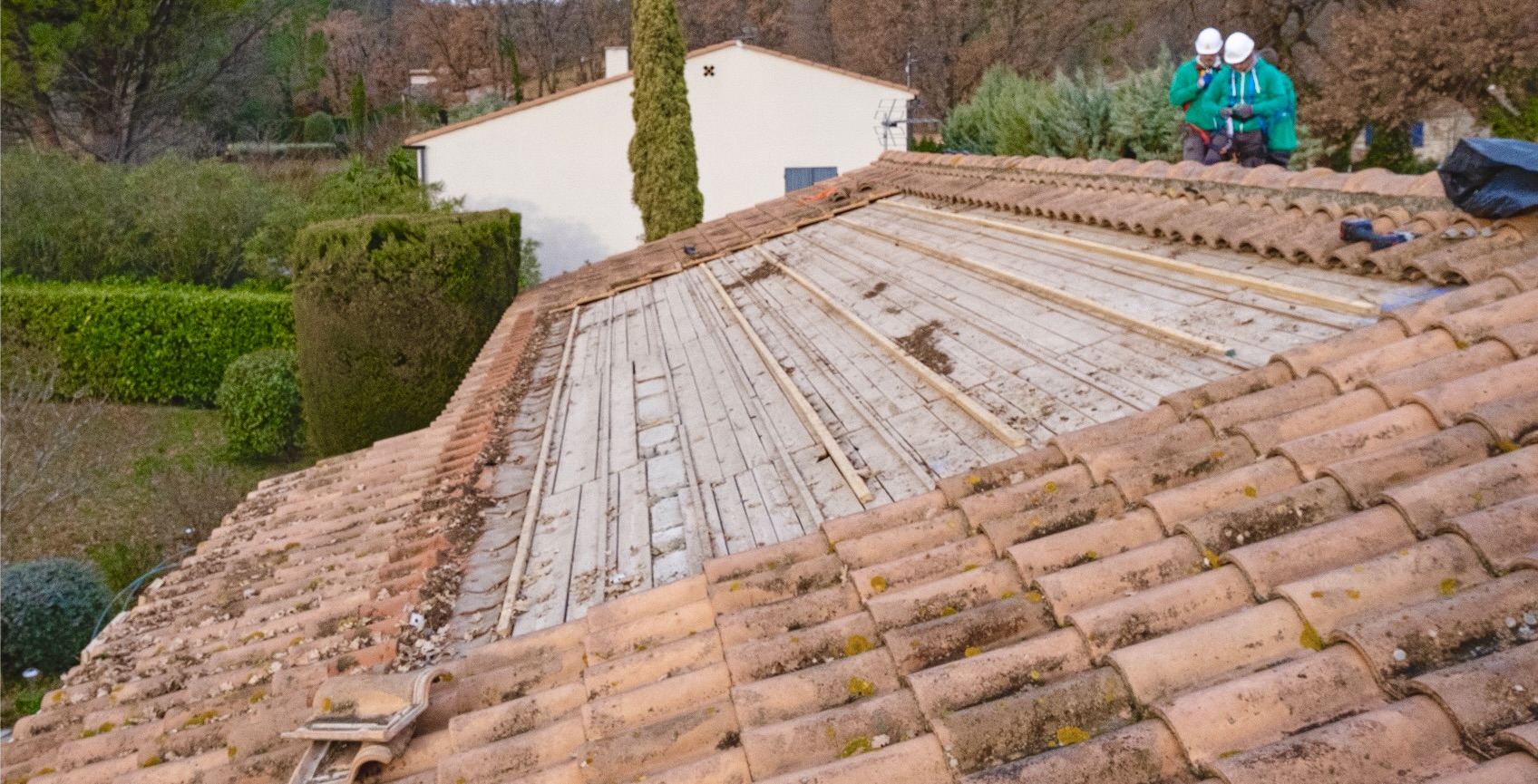 Toiture partiellement detuilee montrant le platelage en bois et liteaux, chantier en cours avec deux ouvriers.