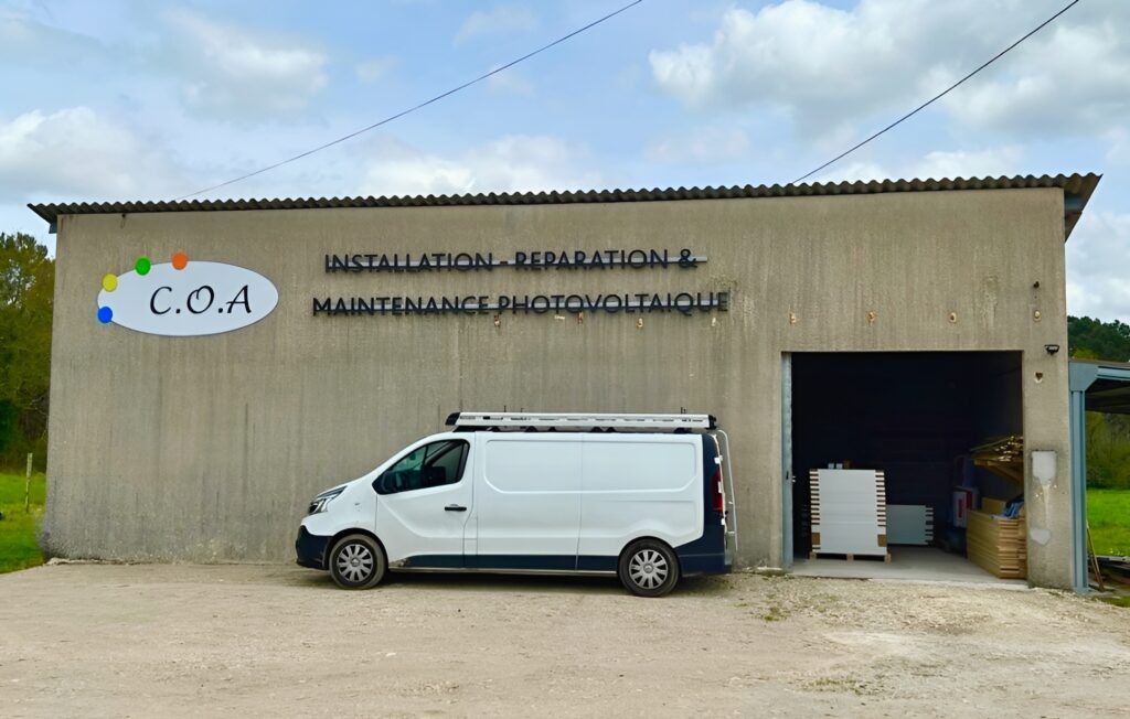 Façade d’un entrepôt gris marquant le logo C.O.A à gauche et l’inscription « Installation, réparation & maintenance photovoltaïque » au centre ; une camionnette blanche est garée devant le bâtiment, dont le grand portail droit est ouvert sur du matériel de panneaux solaires.