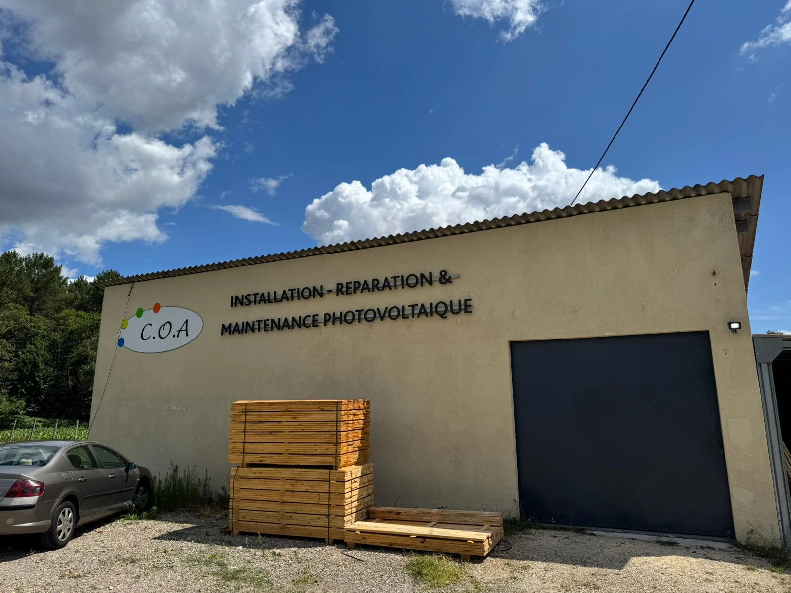 Vue latérale d’un mur beige de l'entrepôt C.O.A de Coimères (proche de Langon) affichant le logo C.O.A et la mention “INSTALLATION – RÉPARATION & MAINTENANCE PHOTOVOLTAÏQUE”, des piles de lames de bois posées devant un grand rideau métallique fermé et une berline grise stationnée sur un sol en gravier sous un ciel partiellement nuageux.