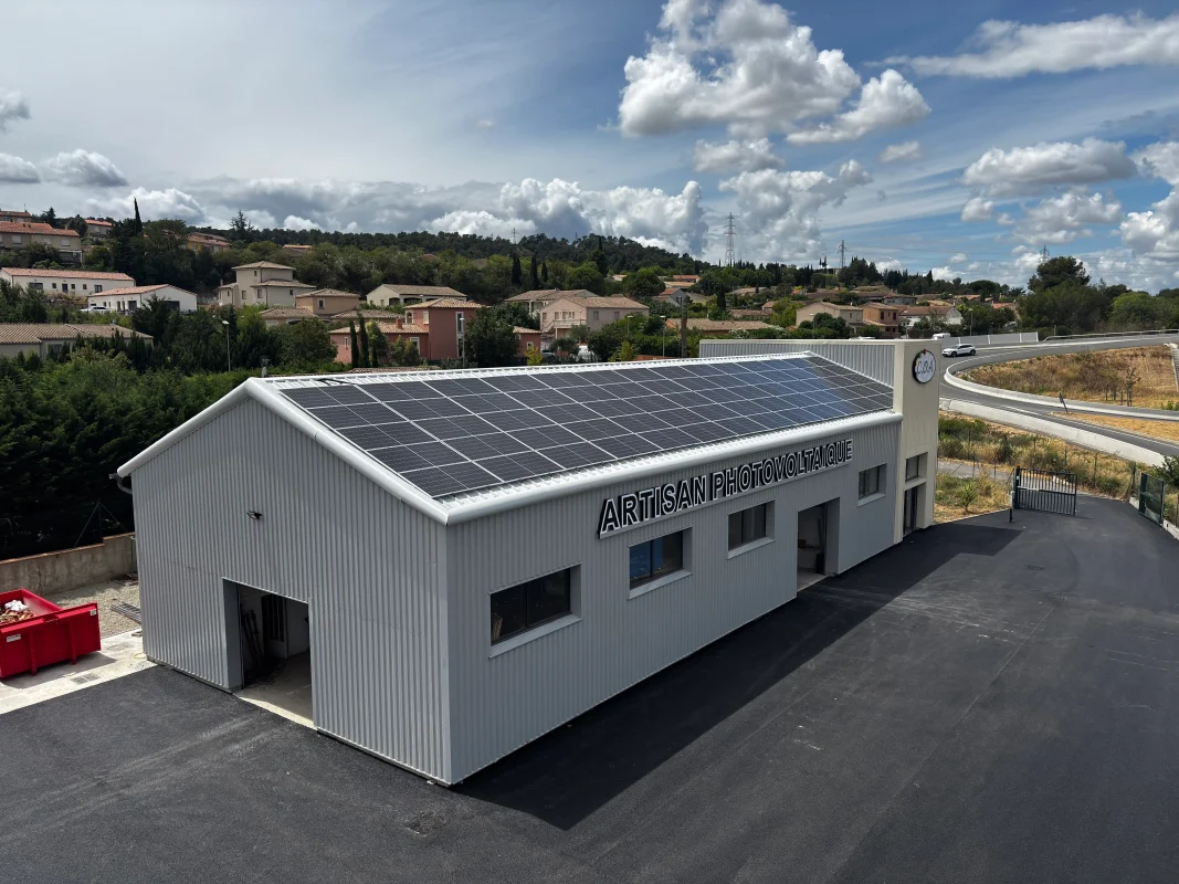 Atelier C.O.A. avec panneaux solaires sur le toit, vaste enrobé noir, accès routier en courbe et collines boisées.